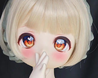 May include: Close-up of a doll's face with large, expressive eyes. The eyes are a mix of orange, brown, and blue, with a heart-shaped reflection. The doll has light blonde hair, rosy cheeks, and a hand near its face.