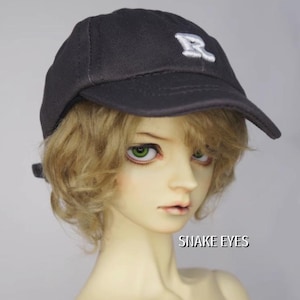 Pode incluir: Boné de beisebol preto com um logotipo "R" branco bordado. O boné é usado por uma boneca com olhos verdes e cabelo loiro. O texto "SNAKE EYES" é visível em branco.