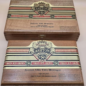 May include: Two wooden cigar boxes, one atop the other, with the Ashton VSG logo prominently displayed. The boxes feature a decorative band with the text "VIRGIN SUN GROWN." The top box is labeled "ASHTON VSG ROBUSTO," and the bottom box is labeled "ASHTON VSG TRES MYSTIQUE."