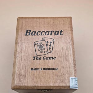 May include: A wooden box with the word "Baccarat" printed on the top. Below the word "Baccarat" are two playing cards and the words "The Game". The box is made in Honduras.