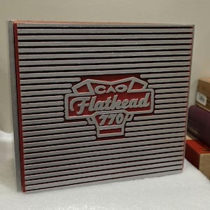 May include: A square, red and silver CAO Flathead 770P box. The box has a red center with the CAO Flathead logo in silver. The box has silver horizontal lines across the top and sides.