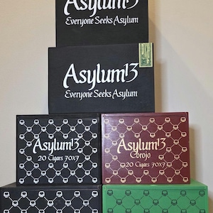May include: A collection of cigar boxes with the "Asylum 13" logo in white script. The boxes are black, burgundy, and green, with skull patterns and text indicating cigar sizes. Some boxes have the text "Everyone Seeks Asylum".