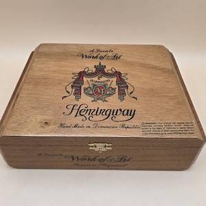 May include: A rectangular wooden cigar box with a hinged lid and a gold-tone metal latch. The lid features the text A Fuente Work of Art, Hemingway, and Hand Made in Dominican Republic, along with a crest logo and a California Proposition 65 warning.