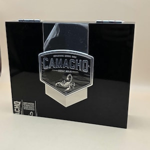 May include: A rectangular black box with a glossy finish featuring a silver shield-shaped emblem on the front. The emblem displays the text CAMACHO, BUILT BOLD, INFAMOUS SINCE 1962, and a scorpion graphic. A small label reads IMPORTED TOBACCO.