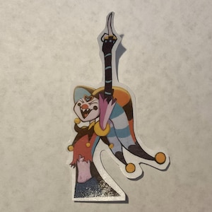 Puede incluir: Una pegatina de un bufón con un gorro de bufón colorido, un traje rojo y amarillo y un cuello de bufón azul y blanco. El bufón sostiene un bastón a rayas blanco y negro en su mano derecha y sonríe con la boca abierta.