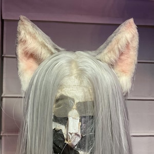 May include: A cosplay wig featuring long, silver-gray hair and attached cat ears. The ears are white with pink inner details. The wig's base is visible, showing a lace front construction. The background is a light purple.