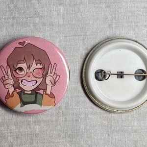May include: A pink button with a cartoon illustration of a person with brown hair, wearing glasses and a green shirt. The person is smiling and giving a peace sign with both hands. The button has a white back with a safety pin.