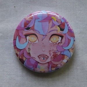 May include: A round button featuring a colourful illustration of a cartoon character with pink and blue hair, yellow eyes, and a pink tongue sticking out. The character is wearing a necklace with colourful beads.