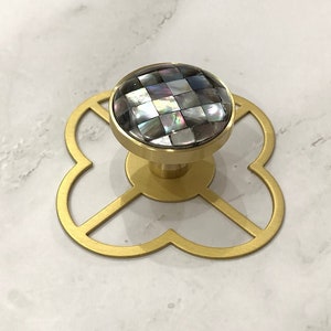 May include: A gold-toned cabinet knob with a decorative, iridescent, mother-of-pearl inlay. The knob has a four-leaf clover-shaped base.