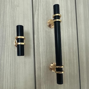 May include: Two black cabinet pulls with gold detailing. One is a short, cylindrical knob, the other a long, vertical bar pull. Both have gold rings and circular base plates. The hardware is mounted on a light wood surface.