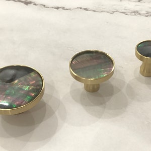 Peut inclure: Trois boutons ronds pour armoire avec une base en métal doré et un incrustation de nacre irisée. Les boutons sont de tailles différentes.