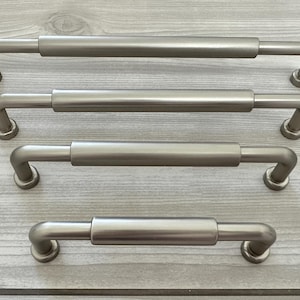 May include: Four brushed nickel cabinet pulls arranged in a vertical stack from longest to shortest. Each handle features a cylindrical centre bar with rounded ends and two mounting posts that extend outward to connect to the cabinet surface.