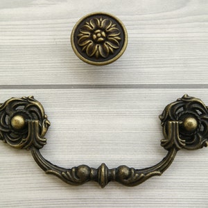 Pode incluir: Puxador de gaveta de latão antigo com detalhes florais ornamentados. O puxador tem uma forma curva e duas maçanetas redondas nas extremidades.
