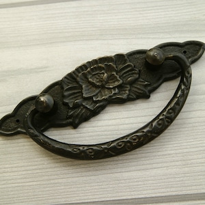 以下が含まれることがあります： 花柄の黒い金属製の引き出しの取っ手。取っ手は湾曲した形をしていて、中央に装飾的な花柄のデザインが施されています。