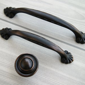 Könnte beinhalten: Zwei schwarze Metall-Schrankgriffe mit verzierten Schnörkeln und einem passenden schwarzen Metallknopf. Die Griffe sind etwa 12,5 cm lang und der Knopf hat einen Durchmesser von 3,8 cm.