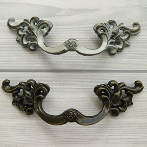 May include: Two ornate drawer pulls, one in white and one in bronze, with a floral design. Both pulls have a curved shape and a rose-shaped accent in the centre.