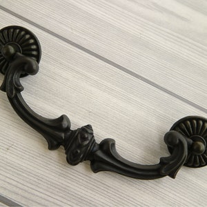 May include: Black metal drawer pull with a decorative, ornate design. The pull has a rounded, fluted knob on each end.