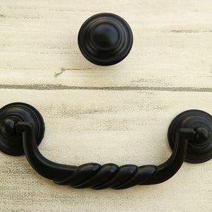 以下が含まれることがあります： ねじれたデザインの黒いキャビネット金具。プルハンドルは湾曲した形状で、両端に丸いノブが付いています。