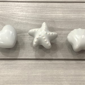 May include: Three white ceramic drawer pulls in the shapes of a heart, a starfish, and a seashell.