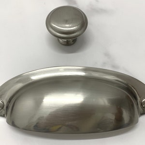 Peut inclure: Un ensemble de deux poignées de meuble en nickel brossé. La plus grande pièce est une poignée de tasse de 7,6 cm avec un bord arrondi. La plus petite pièce est un bouton de 2,5 cm avec un dessus arrondi.