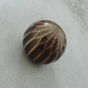 May include: A spherical wooden object with a polished finish. The wood grain features a pattern of light and dark brown concentric lines, creating a textured appearance. The object is set against a neutral background.