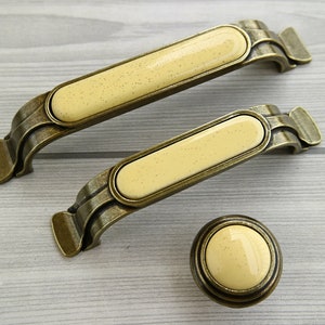 May include: Two antique gold cabinet pulls with cream-colored ceramic inserts and one matching knob. The pulls have a decorative, ornate design.