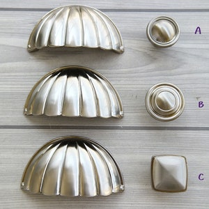 Puede incluir: Cuatro pomos y tiradores de armario en acabado de níquel cepillado. Los pomos son redondos y los tiradores tienen forma de concha de vieira. Los pomos están etiquetados como A, B y C, y los tiradores como D.