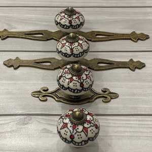 以下が含まれることがあります： 白い背景に赤と黒の花柄が描かれたセラミック製のノブが4つ。各ノブは金色の装飾的な金属プレートに取り付けられています。