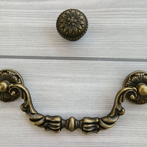 May include: A decorative brass drawer pull with ornate details. The pull has a curved shape with two round knobs on either end. The knobs have a floral design.