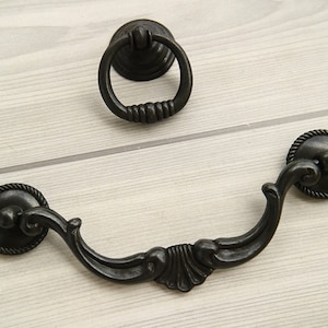 May include: Two black metal cabinet pulls. One is a ring pull and the other is a curved pull with a decorative design.