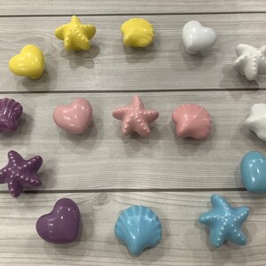 May include: Assortment of ceramic drawer pulls in various shapes and colors, including seashells, starfish, and hearts. The pulls are all in pastel colors, including pink, blue, yellow, and purple.