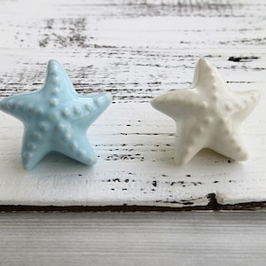 May include: Two ceramic starfish drawer pulls, one light blue and one white, on a whitewashed wood background.