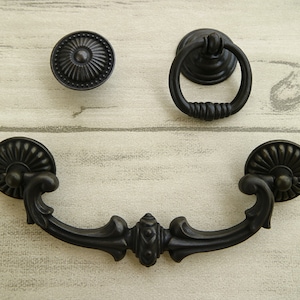 Puede incluir: Tres tiradores de armario de metal negro con diseños ornamentados. Uno es un pomo redondo, otro es un tirador de anillo y el último es un tirador decorativo con una manija curva.