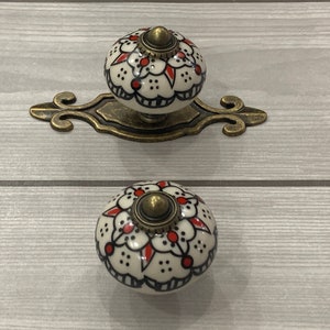 以下が含まれることがあります： 白地に赤と黒の花柄が描かれたセラミック製のノブが2つ。ノブは真鍮仕上げで、真鍮製のプレートに取り付けられています。