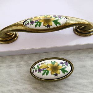 May include: A pair of oval-shaped cabinet knobs with a floral design. The knobs are white with yellow and purple flowers and green leaves. The knobs are attached to a gold-colored metal base.