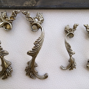 May include: Three ornate metal drawer pulls with a leaf design. The pulls are silver in colour and have a decorative scrollwork pattern.