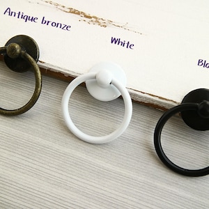 Op de afbeelding: Drie ringgrepen in verschillende kleuren, antiek brons, wit en zwart. De ringgrepen liggen op een wit houten oppervlak.