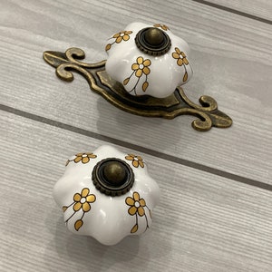 May include: Two white ceramic cabinet knobs with a floral design in gold and brown. The knobs are mounted on a decorative bronze-coloured backplate. The knobs are designed for furniture and home decor.