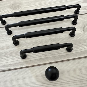 May include: A collection of four matte black cabinet pulls of varying lengths, each featuring a cylindrical bar design with rounded ends and circular mounting bases, accompanied by a single matching matte black spherical cabinet knob.