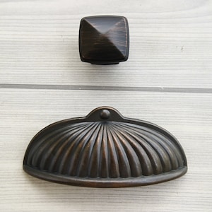 Pode incluir: Duas puxadores de armário em acabamento bronze oleado. Uma é um botão quadrado com um centro ligeiramente elevado. A outra é uma puxador curvo com um design nervurado.