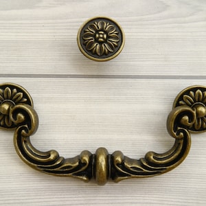 May include: Antique brass cabinet pull with ornate floral details. The pull has a curved shape and two decorative knobs on either end.