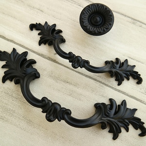 May include: Two black ornate metal drawer pulls with a decorative floral design. The pulls are in the shape of a curved handle with a leaf-like design on each end.