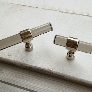 May include: Two clear glass cabinet knobs with a silver finish. The knobs are in the shape of a T with a round base.