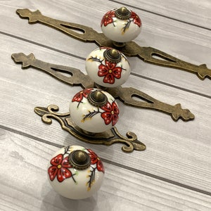 May include: Four white ceramic cabinet knobs with red floral designs and gold accents. Each knob is attached to a gold metal base.