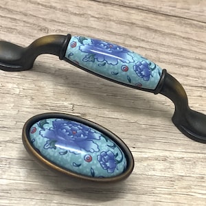 May include: Two antique-style cabinet pulls. The pulls have a dark bronze finish with a ceramic center. The ceramic center features a blue floral design on a light blue background. One pull is a handle, and the other is an oval knob.