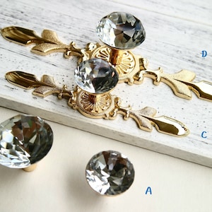 May include: Four crystal cabinet knobs with gold metal bases. The knobs are in the shape of a diamond and have a faceted design. The bases are ornate and have a leaf design.