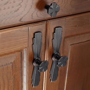 Peut inclure: Deux poignées de porte de placard en métal noir avec un design en forme d'éventail décoratif et un bouton en forme de croix. Les poignées sont montées sur une porte de placard en bois.