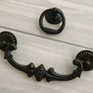 以下が含まれることがあります： 黒い金属製のキャビネットプル2個。1つはリングプル、もう1つは装飾的なディテールが施された湾曲したプルです。
