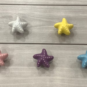 May include: Five ceramic starfish drawer pulls in different colours: white, yellow, pink, purple, and blue. The starfish are all the same size and have a raised, textured surface.
