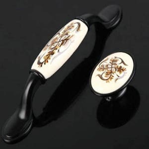 Pode incluir: Um par de botões de armário de cerâmica preta e branca com um design floral. Os botões têm uma forma curva e uma base redonda.